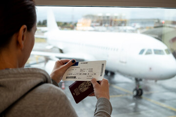 A woman at an international airport, holding a passport and airline tickets in her hand, and looking at her plane through the window. Flight delay. Selective focus.