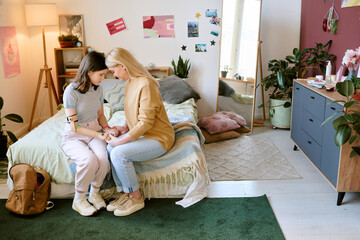 Two young women sitting on bed in cozy bedroom, offering support and comfort during emotional...