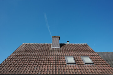 A close-up view of a sloped rooftop covered in weathered red tiles, featuring two rectangular skylights and a central chimney.