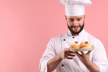 Happy confectioner in uniform holding profiteroles with strawberries on pink background