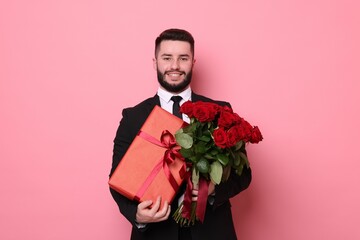 Happy man with bouquet of red roses and gift box on pink background