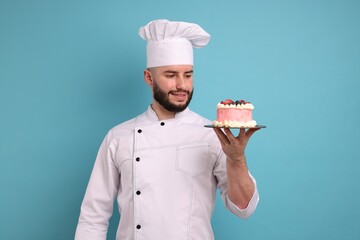 Happy confectioner in uniform holding cake with berries on light blue background
