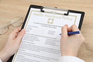 Doctor filling out health insurance policy at wooden table, closeup