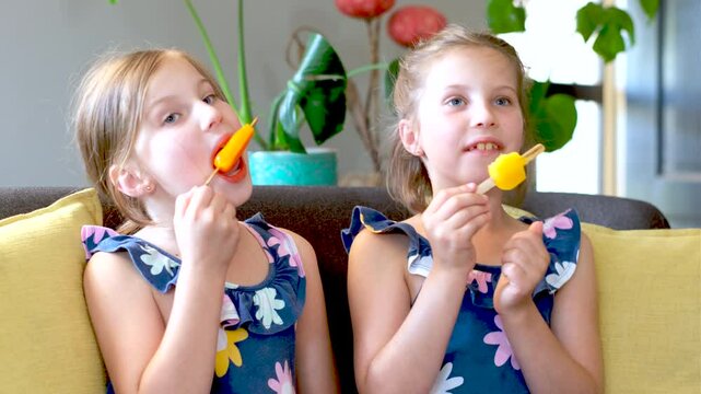 Frontal view of animated fraternal twin European girls eating orange ice pops