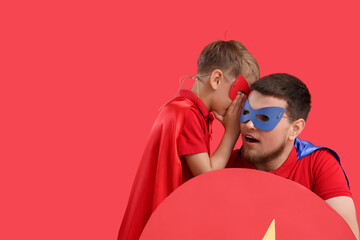 Little boy dressed as superhero whispering in his father's ear on red background