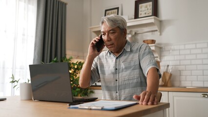 Stressed senior manager talking to colleague by using smart phone and planning marketing strategy. Businessman checking report and calling colleague while sitting at home. Remote working. Myrmidon.