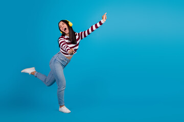 Cheerful young woman in casual attire dancing energetically with headphones against a vibrant blue background