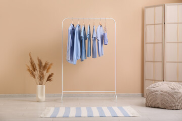 Rack with stylish clothes, pouf and folding screen near beige wall in room