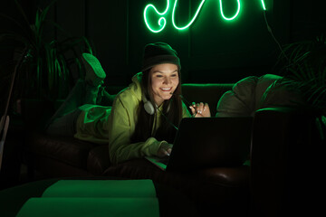 Young woman with headphones sitting on sofa and using laptop in dark living room at night