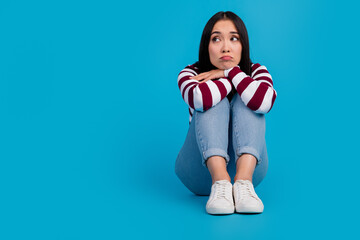 Pensive young woman sitting against a vibrant blue backdrop in trendy casual jeans
