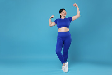 Fototapeta premium Plus size woman in gym clothes showing her biceps on light blue background, space for text
