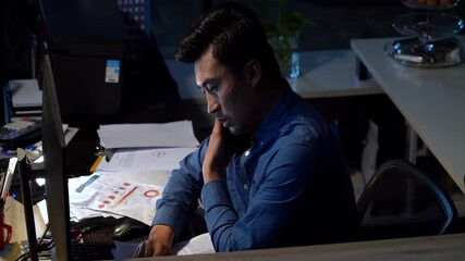 Young businessman feeling overwhelmed by a mountain of paperwork. He is feeling stressed and frustrated as he looks at computer screen