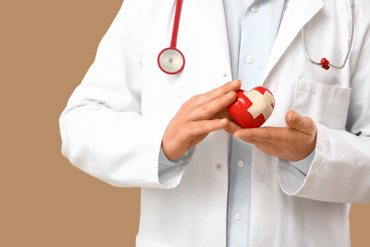 Young male doctor holding red heart with medical patch on beige background, closeup. World Health Day