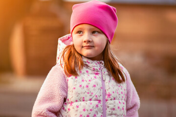 A little girl with light brown hair and fair skin wears a pink beanie and a white floral jacket....