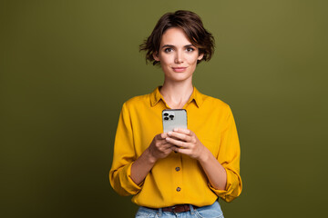 Confident young brunette woman holding smartphone wearing yellow stylish shirt against green...