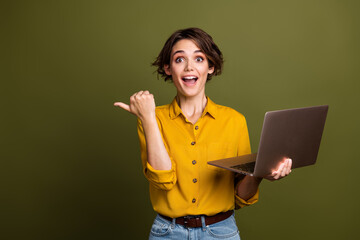 Smiling young woman holding a laptop and pointing to the side against a green background,...