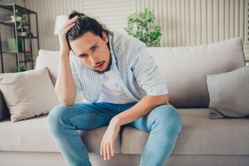 Obraz premium Young man casually dressed sitting on a sofa in a bright modern living room, looking thoughtful, relaxed and deep in contemplation