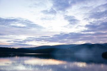 Tuyen Lam Lake in Da Lat at sunset, with mist rising above the calm water. The soft light reflects off the lake, surrounded by rolling hills and forests, creating a serene and dreamy atmosphere.