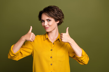 Confident young woman in yellow shirt showing thumbs-up gesture on khaki background symbolizing...