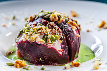 Beetroot with cottage cheese cream, nuts and pumpkin seeds on green spinach sauce is served on a white plate. Restaurant serving of a healthy dish. Close-up.