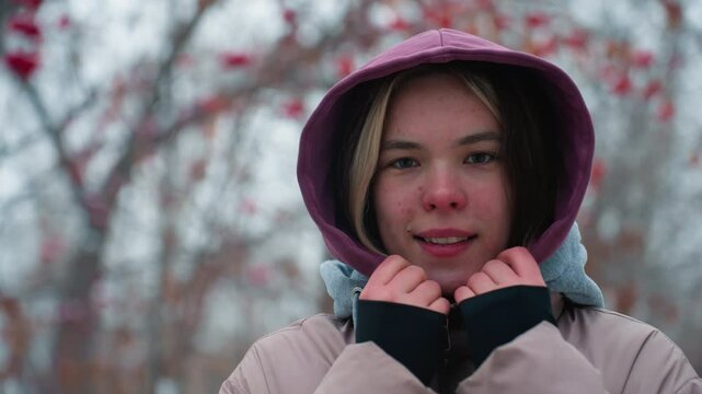 close up of young lady smiling slightly while holding her hoodie collar, looking at camera with subtle smile, winter jacket, outdoor setting with blurred trees in background