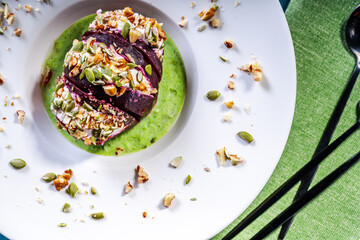 Beetroot with cottage cheese cream, nuts and pumpkin seeds on green spinach sauce served on a white plate with a napkin and cutlery. Restaurant serving of a healthy dish. Top view