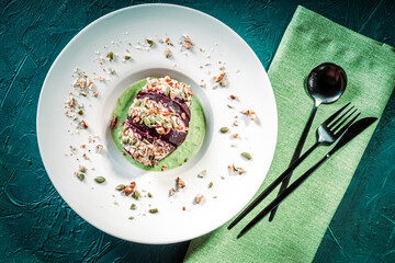 Beetroot with cottage cheese cream, nuts and pumpkin seeds on green spinach sauce served on a white plate with a napkin and cutlery. Restaurant serving of a healthy dish. Top view