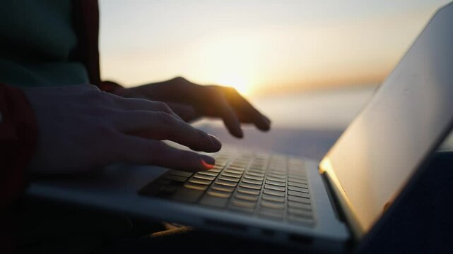 Freelancer typing on laptop keyboard during golden hour, creating peaceful work environment with ocean and sunset backdrop, embodying digital nomad lifestyle and remote work freedom