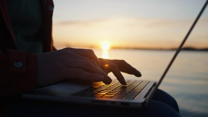 Man entrepreneur working on laptop near scenic lake during golden sunset, typing intently and closing device after completing professional task, embodying digital nomad lifestyle