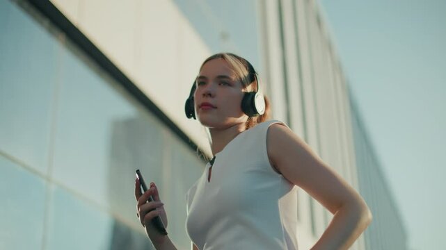 Upper angle view of young girl dancing with headset on and phone in right hand, eyes closed in rhythm, surrounded by modern glass building reflecting sky in urban environment
