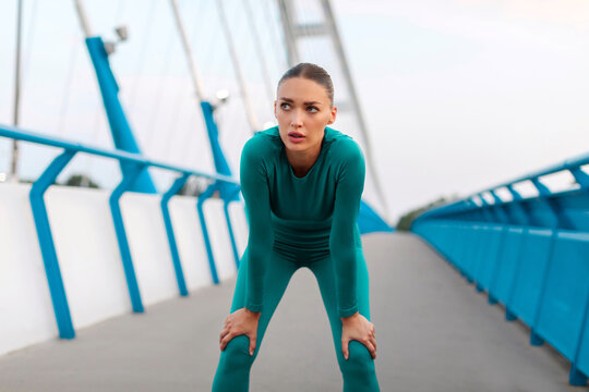 Woman runner in sports outfit feeling tired, resting after running on the bridge in the city, taking break and breathing