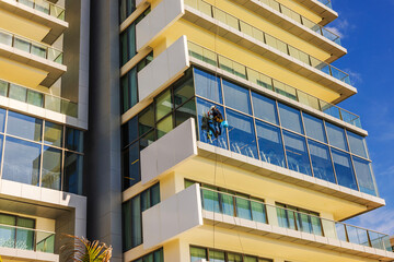 Obraz premium Worker suspended on ropes cleaning windows of modern hotel building with blue sky in Caribbean resort. Mexico. Cancun. 