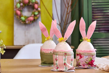 three handmade Easter bunnies made of eggs with pink ears stand on a table against a green wall