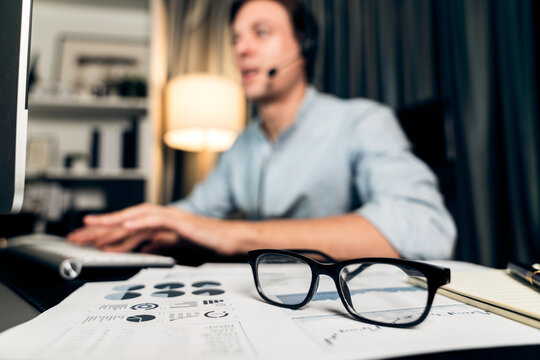 Call center consulting with customers on headphones on monitor in business paragraph with typing on pc with dynamic data marketing analysis planning at modern home office at night time. Pecuniary.