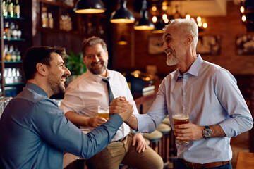 Cheerful businessmen greeting with manly handshake while drinking beer in pub.