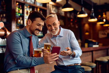 Happy businessman showing something on mobile phone to his colleague while drinking beer in bar.