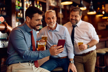 Smiling entrepreneur and his coworkers using cell phone while drinking beer in pub.