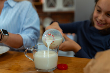 Happy girl pouring milk from a plastic bottle into a glass mug at the breakfast table, with family members sitting nearby, enjoying a healthy and nutritious start to the day