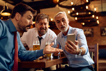 Male colleagues watching sports game on mobile phone and drinking beer in pub.