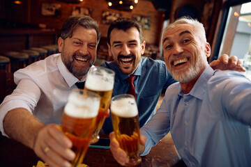 Cheerful businessmen taking selfie while drinking beer in pub and looking at camera..