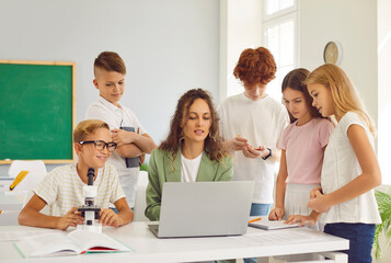 School teacher and students learning, pupils around desk watching laptop in classroom. Female tutor and smart group gathering around table during lesson at school, watching educational video online 