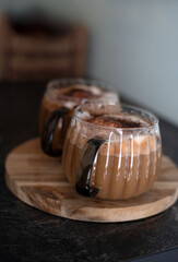 two frothy coffee beverages in glass mugs with dark handles on a rustic wooden board, set against a...