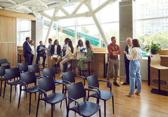 Full length photo of a group of diverse business people men and women chatting after a meeting. Company employees team or group of staff talking in office or big conference room with empty chairs.