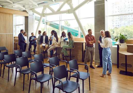 Full length photo of a group of diverse business people men and women chatting after a meeting. Company employees team or group of staff talking in office or big conference room with empty chairs.