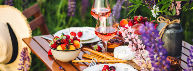 Banner. Two glasses of rose wine, flowers in a vase, silverware, fruits. Sunset, golden hour, atmospheric candlelight. Romantic table decor for a loving couple on blooming meadow with purple lupines