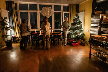 An elderly man and two women preparing for a Christmas dinner in a cozy home at night. A decorated Christmas tree with gifts is visible, with candles lighting the festive table setting.