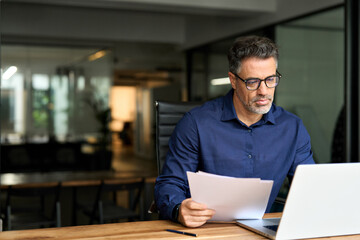 Middle aged professional business man using laptop computer reading document at work. Busy mature male account bank manager executive working on laptop checking financial report in office. Copy space.
