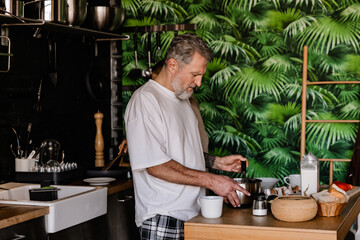 A White elder man in a white T-shirt and plaid pants prepares breakfast in a cozy kitchen, stirring...