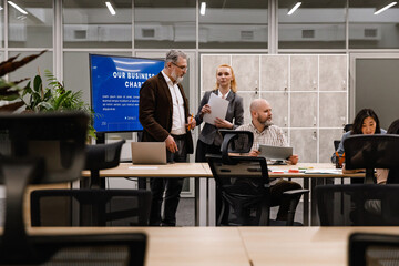 A senior man in a blazer and a young woman in a business suit stand near a screen during a corporate presentation. A group of professionals, including a bald man and an Asian woman, discuss documents.
