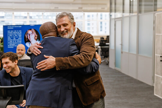 A White elder man in a brown blazer embraces a Black bald man with an earring in a modern office, while colleagues are engaged in conversations and laughter, fostering a friendly workspace atmosphere.